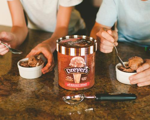 Two people enjoy bowls of Rocky Road ice cream from a Dreyer's container, with spoons in hand on a kitchen counter.
