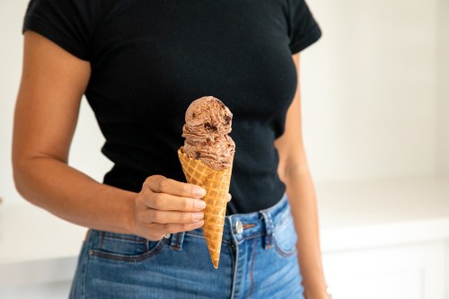 A person holding a chocolate ice cream cone with a waffle cone, wearing a black shirt and blue jeans.