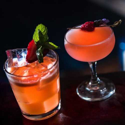 Two colorful cocktails on a bar: one in a rocks glass with ice and mint, the other in a coupe glass with a garnish.