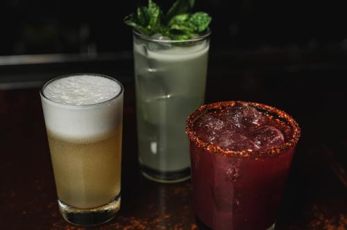 Three cocktails on a bar: a frothy beer, a minty green drink, and a dark red cocktail with a spicy rim.