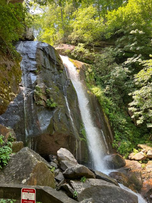 A waterfall cascading down rocky cliffs, surrounded by lush green trees and vegetation. A warning sign is visible nearby.