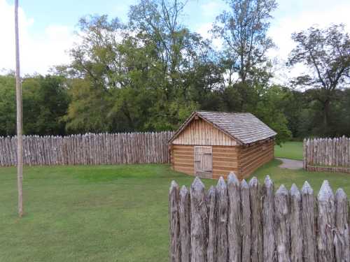 A wooden cabin surrounded by a tall wooden fence, set in a grassy area with trees in the background.