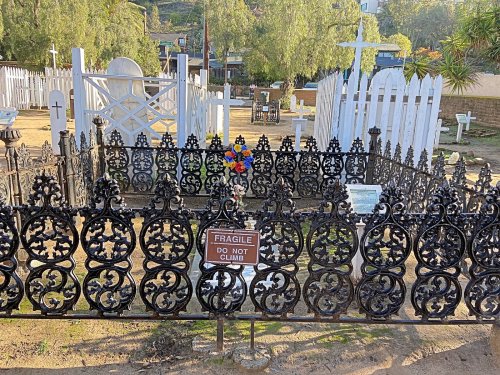 A decorative iron fence surrounds a grave site with flowers and a sign that reads "Fragile, Do Not Climb."