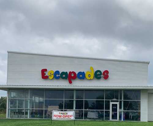 Colorful sign reading "Escapades" on a storefront with a "Now Open!" banner in front. Cloudy sky above.