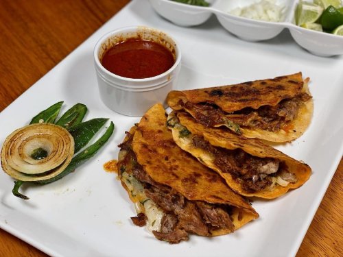 Three crispy tacos filled with meat, served with a side of red sauce, grilled onions, and jalapeños on a white plate.