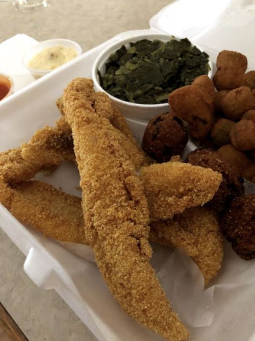A takeout container with fried fish fillets, collard greens, fried okra, and a small cup of dipping sauce.