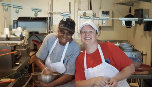 Two women in a kitchen, one mixing ingredients in a bowl, both smiling and wearing aprons and caps.