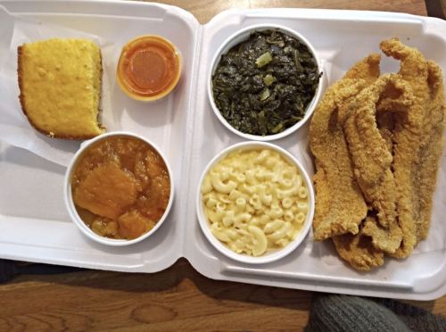 A takeout container with fried catfish, macaroni and cheese, collard greens, sweet potato casserole, and a sauce.