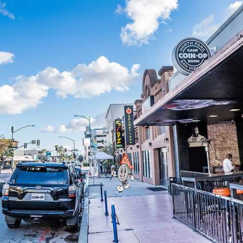 A street view featuring a coin-op game room, parked cars, and a blue sky with clouds.