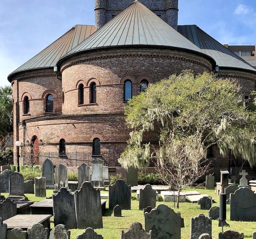 Historic brick building with a round roof, surrounded by a cemetery featuring weathered gravestones and lush greenery.