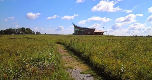 A winding path leads through tall grass to a modern building under a blue sky with fluffy clouds.