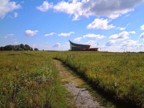 A modern building with a curved roof sits on a grassy field under a blue sky with fluffy clouds.