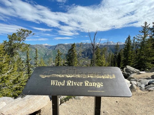 Sign for the Wind River Range with a scenic mountain view and blue sky in the background, surrounded by trees.