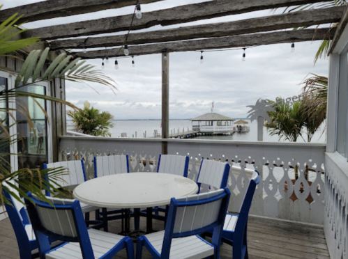 A coastal deck with a round table and blue chairs, overlooking a calm water view and a distant pier.