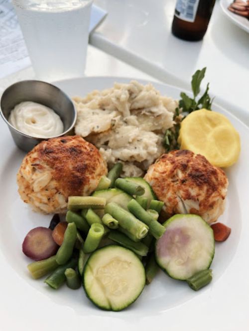 A plate of crab cakes, mashed potatoes, mixed vegetables, and a lemon wedge, served with a small cup of sauce.