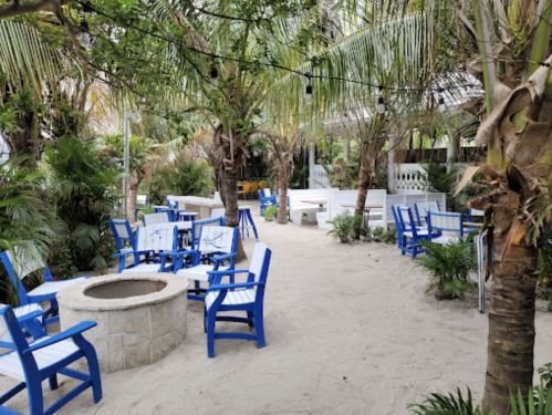 A sandy outdoor area with blue chairs, a stone fire pit, and lush tropical plants.