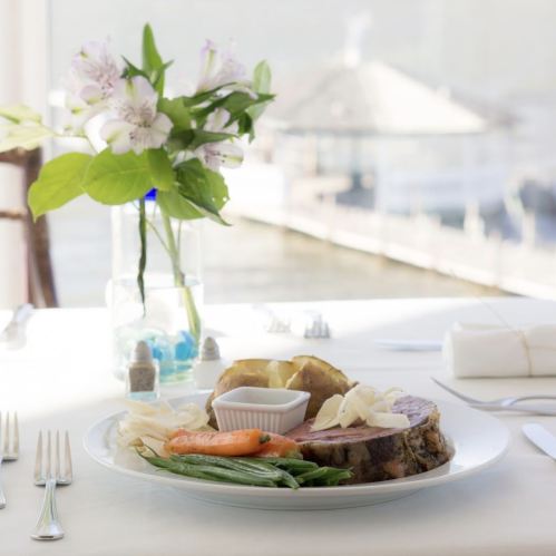 A plate of sliced roast beef with green beans, carrots, and a baked potato, set on a table with a flower vase.