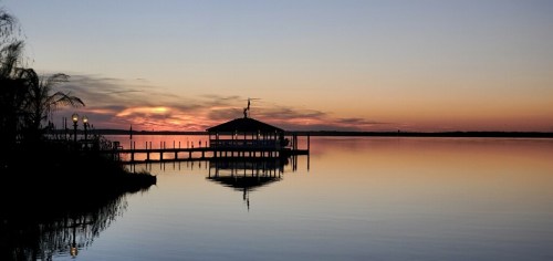 A serene sunset over a calm lake, with a silhouetted dock and reflections on the water.