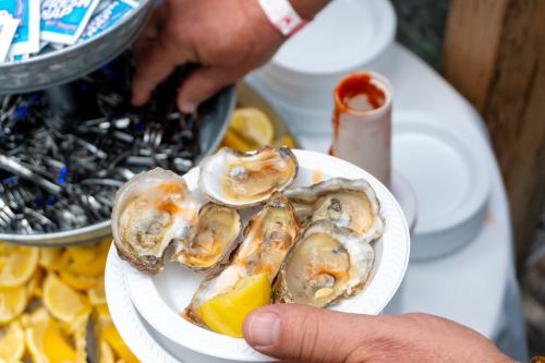 A hand holding a plate of oysters with a lemon wedge, next to a bowl of cocktail sauce and a stack of plates.