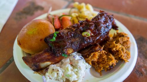 A plate of barbecue ribs, pulled pork, coleslaw, macaroni, a roll, and pickled vegetables.
