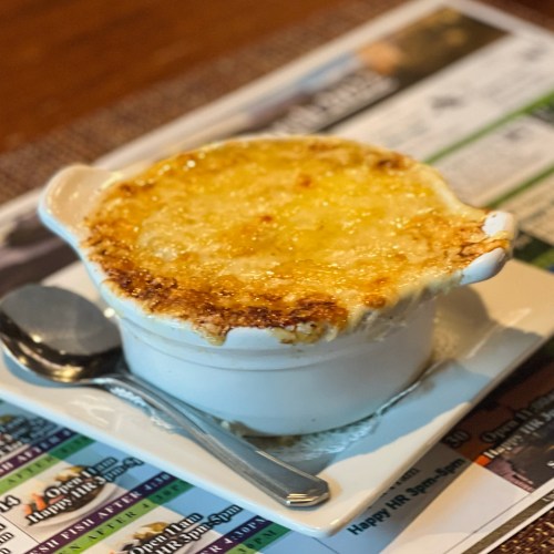 A baked dish with a golden, cheesy crust in a white bowl, served on a plate with a spoon beside it.