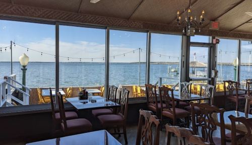 Interior of a restaurant with wooden tables and chairs, large windows overlooking a calm waterfront with string lights.