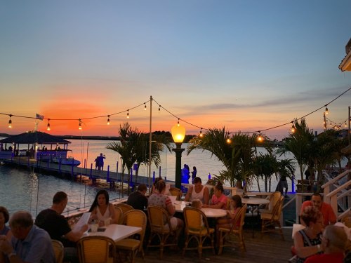 A vibrant sunset over a waterfront dining area, with people enjoying meals and string lights illuminating the scene.