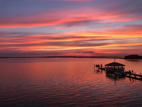 A vibrant sunset over calm waters, with colorful clouds reflecting on the surface and a dock in the foreground.