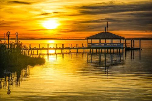 A serene sunset over a calm lake, featuring a dock and a gazebo silhouetted against golden skies.