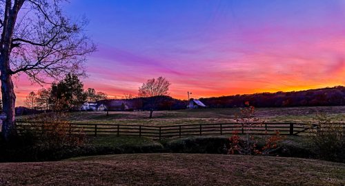 A serene landscape at sunset, featuring a colorful sky, a wooden fence, and trees in a rural setting.