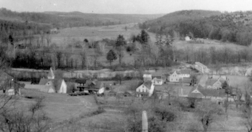 A black-and-white landscape showing a small village surrounded by fields and hills in the background.