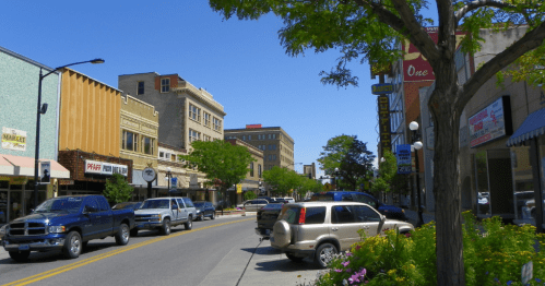 A sunny street scene with shops, trees, and parked cars in a small town. Bright blue sky overhead.