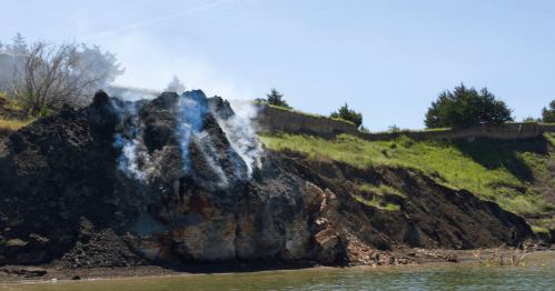 Smoke rises from a rocky hillside near a body of water, with green vegetation in the background under a clear blue sky.