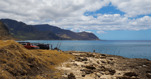A rocky coastline with fishing boats, mountains in the background, and a clear blue sky with scattered clouds.