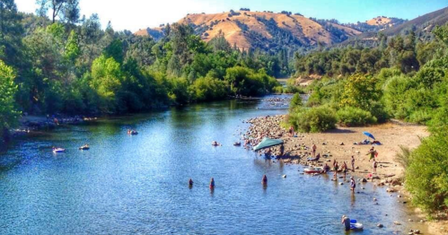 A scenic river surrounded by greenery, with people enjoying the water and sun on a sandy shore.
