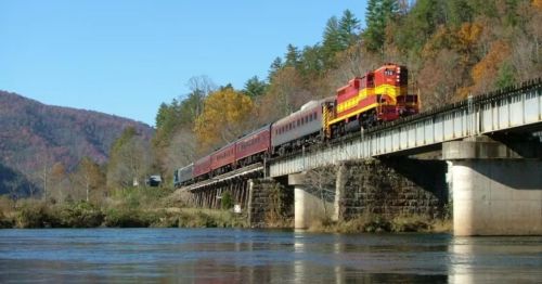A colorful train crosses a bridge over a river, surrounded by autumn trees and mountains under a clear blue sky.