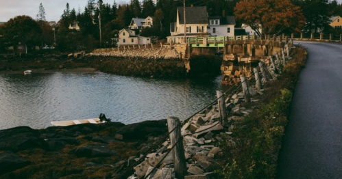 A serene coastal scene with a rocky shore, a small boat, and quaint houses in the background under a clear sky.