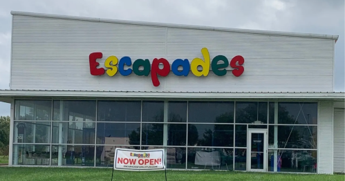 Colorful sign for "Escapades" on a modern building, with a banner stating "NOW OPEN!" in front.