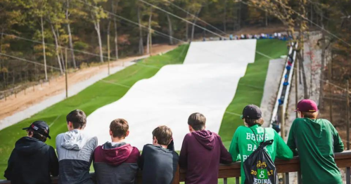 A group of boys watches a ski slope surrounded by trees, with people skiing in the distance.