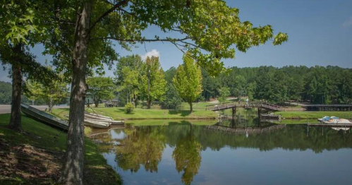 A serene lake scene with trees, a wooden bridge, and boats reflected in the calm water under a clear blue sky.