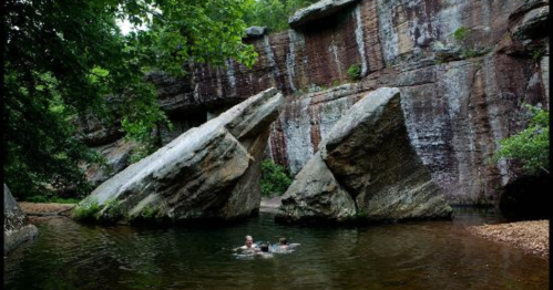Two people swim in a serene pool surrounded by large rocks and lush greenery.