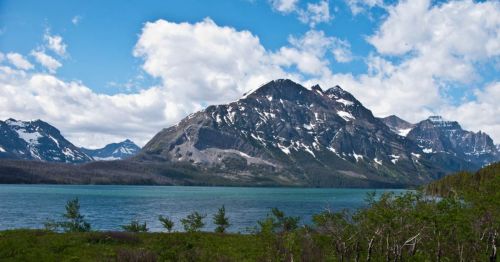 A scenic view of a mountain range with snow-capped peaks, overlooking a calm lake under a partly cloudy sky.