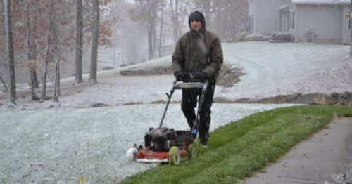 A person in a hooded jacket mows a snowy lawn, surrounded by trees and a house in the background.
