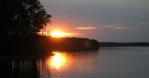 A serene sunset over a calm lake, with silhouettes of trees along the shore reflecting in the water.
