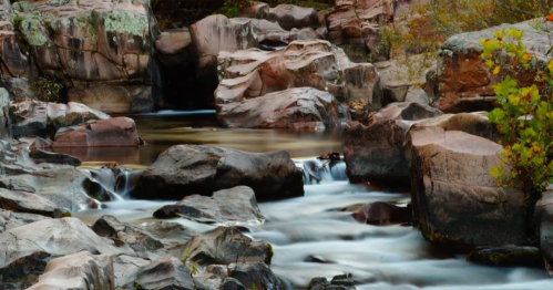 A serene river flows over smooth rocks, surrounded by lush greenery and rugged terrain.