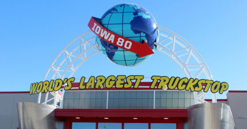 Sign for Iowa 80, the world's largest truck stop, featuring a globe and an arrow, against a clear blue sky.