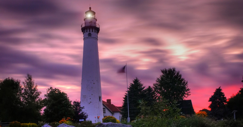 A tall lighthouse stands against a colorful sunset, surrounded by trees and a flag, with dramatic clouds in the sky.
