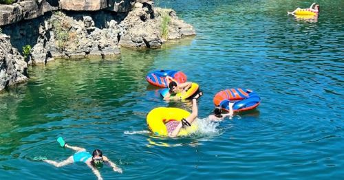 A group of people enjoying a sunny day on colorful inner tubes in a clear blue lake. One person swims nearby.