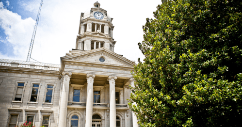 Historic building with a clock tower, columns, and a large tree in the foreground against a bright blue sky.