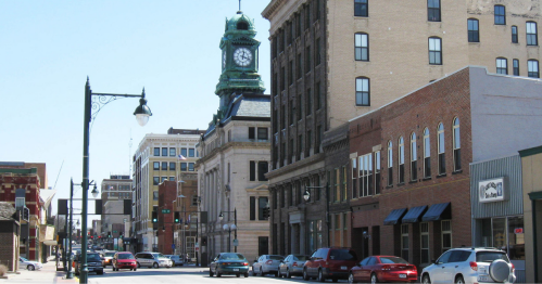 A city street lined with historic buildings, featuring a clock tower and parked cars under a clear blue sky.